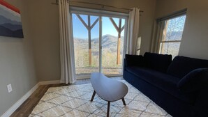 Un salón con un sofá azul oscuro, una silla blanca y una alfombra estampada. Hay un ventanal con cortinas que ofrece vistas a un paisaje montañoso.