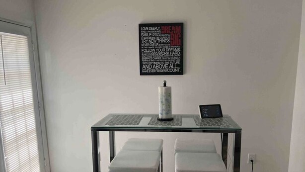Moderni ruokailutila, jossa on lasipöytä, kaksi valkoista tuolia ja kannettava tietokone. Seinällä on näkyvillä inspiroiva sitaatti.