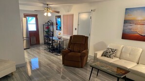 A modern living room with a brown recliner, a white sofa, and a glass coffee table. There is a dining area with a black metal table and chairs, and a kitchen visible in the background. - 2 Bedroom Home 20 Minutes From Everything! (North Las Vegas)