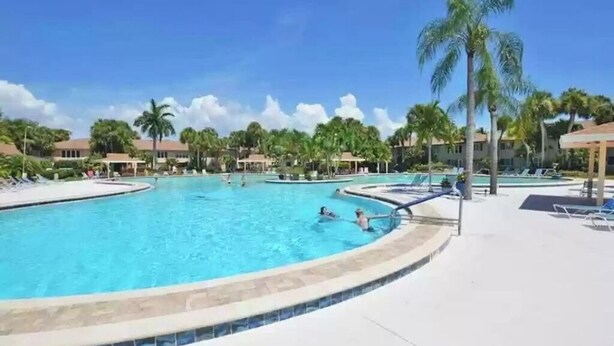 A large outdoor swimming pool surrounded by palm trees and lounge chairs, with a clear blue sky above.