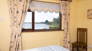 Een slaapkamer met een bed, een stoel, een raam met gordijnen en uitzicht op een meer.