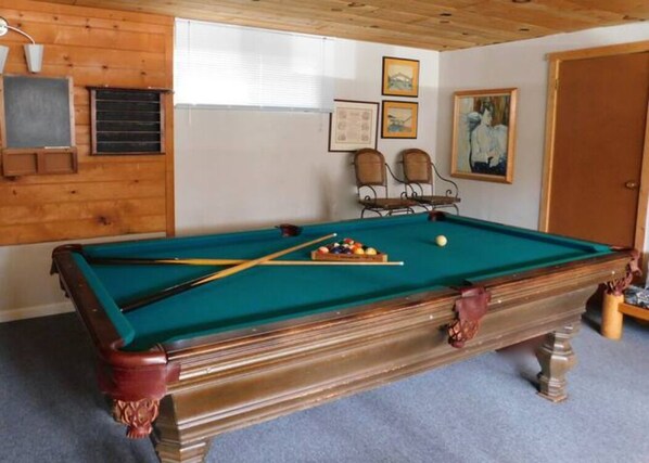 Ein Billardtisch mit Kugeln und Spielstöcken, Holzwände, zwei gerahmte Bilder und eine Tür.