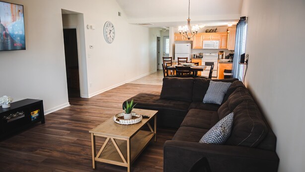 A modern living room with a dark brown sofa, a wooden coffee table, and a TV on the left. A kitchen is visible in the background with wooden cabinets and a chandelier.