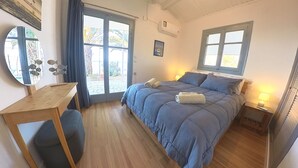 A bedroom with a bed, a desk, and a chair. There is a window with curtains, a picture on the wall, and an air conditioning unit.