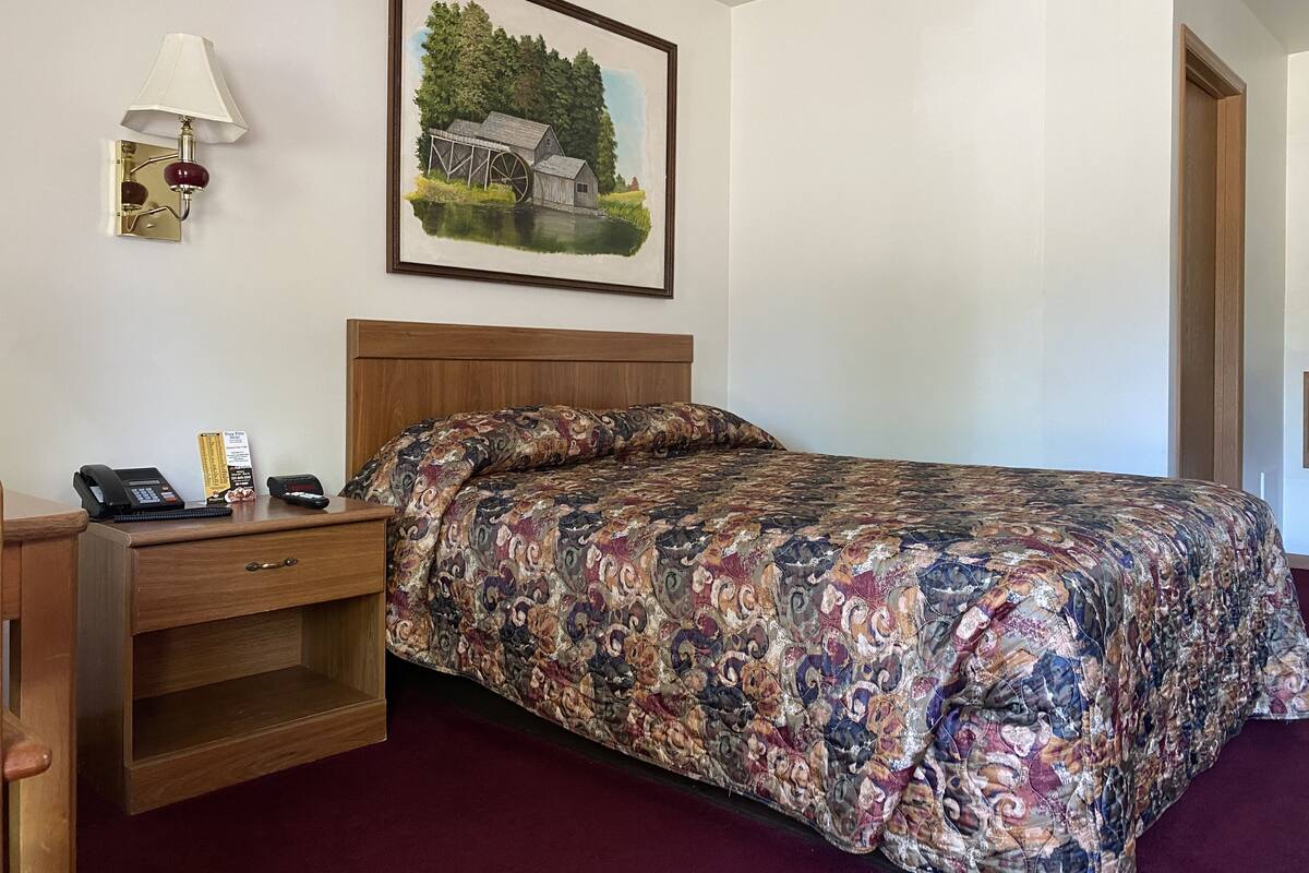 A hotel room with a bed, a desk with a phone and a notepad, a lamp, and a painting of a rural scene.