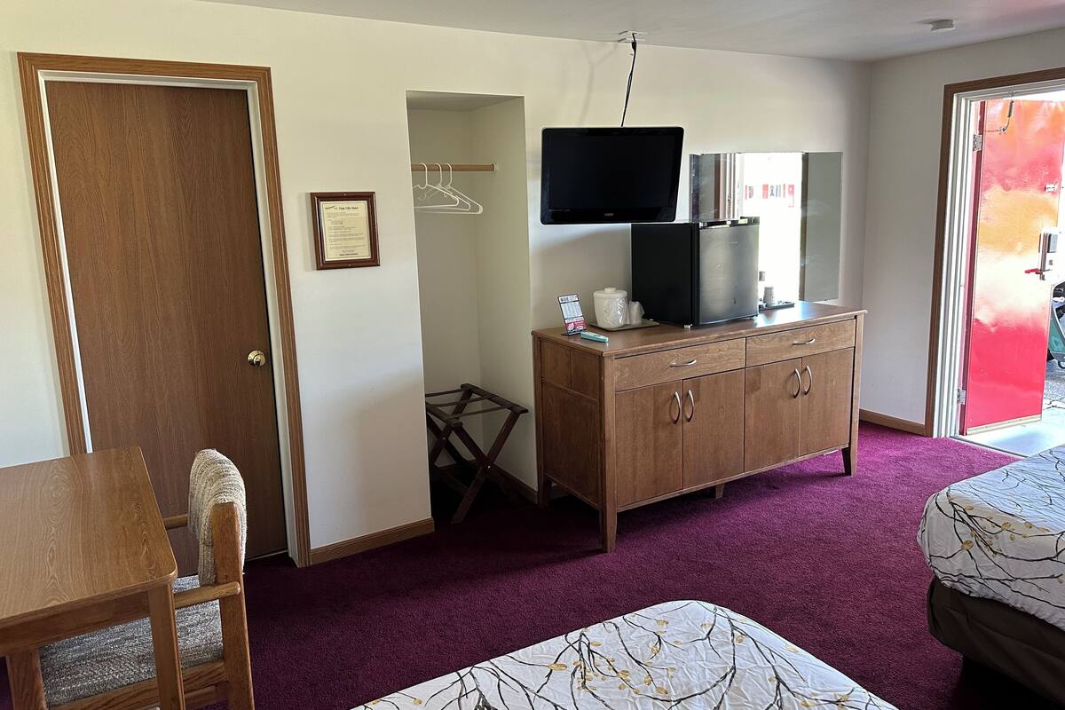 A hotel room with a wooden dresser, a television, a small table with a calendar, a chair, a bed with a patterned bedspread, and a door leading to another room.