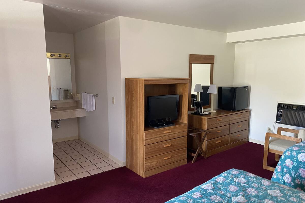 A hotel room with a wooden dresser, a television, a desk with a computer, a mirror, and a bathroom visible through an open door.