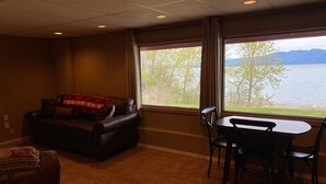 A living room with a sofa, a table, and chairs, a large window with a view of a lake and mountains.