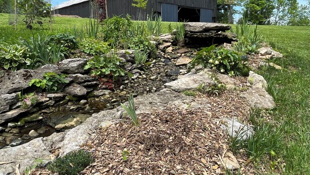 Un granero rústico con una amplia puerta abierta, rodeado de un jardín cuidado con diversas plantas y un pequeño arroyo.