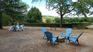 An outdoor seating area with plastic chairs and a small table, surrounded by trees and a hedge.
