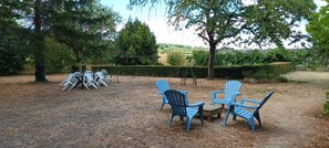 An outdoor seating area with plastic chairs and a small table, surrounded by trees and a hedge.