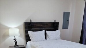 Quarto com piso de madeira, cama com roupa de cama branca, mesa de cabeceira com luminária e luminária fixada na parede.