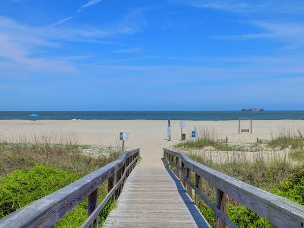 A wooden boardwalk leading to a beach with a ship in the distance.