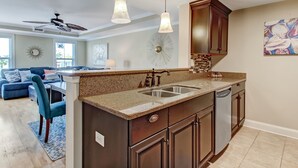 A modern kitchen with dark wood cabinets, a granite countertop, and stainless steel appliances. There is a dining area with a blue sofa and a ceiling fan. - Tybee Getaway Just Steps to the Beach! (Tybee Island)