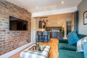 Un salon moderne avec un mur en briques, un téléviseur à écran plat, un canapé vert et un pouf à motifs écossais.