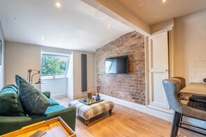 A modern living room with a brick wall, a flat-screen TV, a green sofa, a plaid ottoman, and a wooden coffee table.