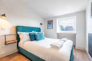 A neatly made bed with white and teal bedding, a bedside table with a lamp, and a window with a view of trees.