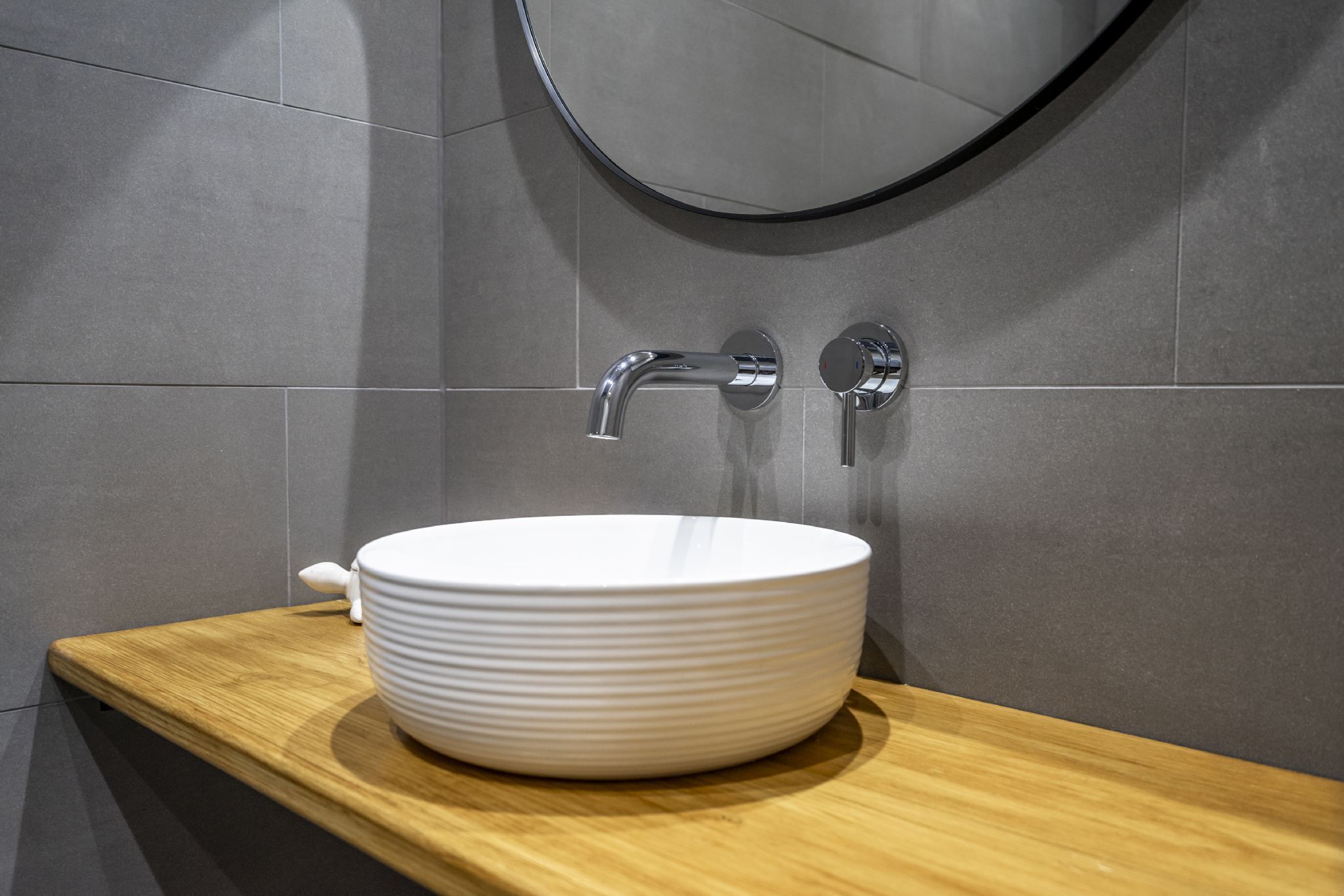 A modern bathroom with a white ceramic sink, a wooden countertop, and a round mirror.