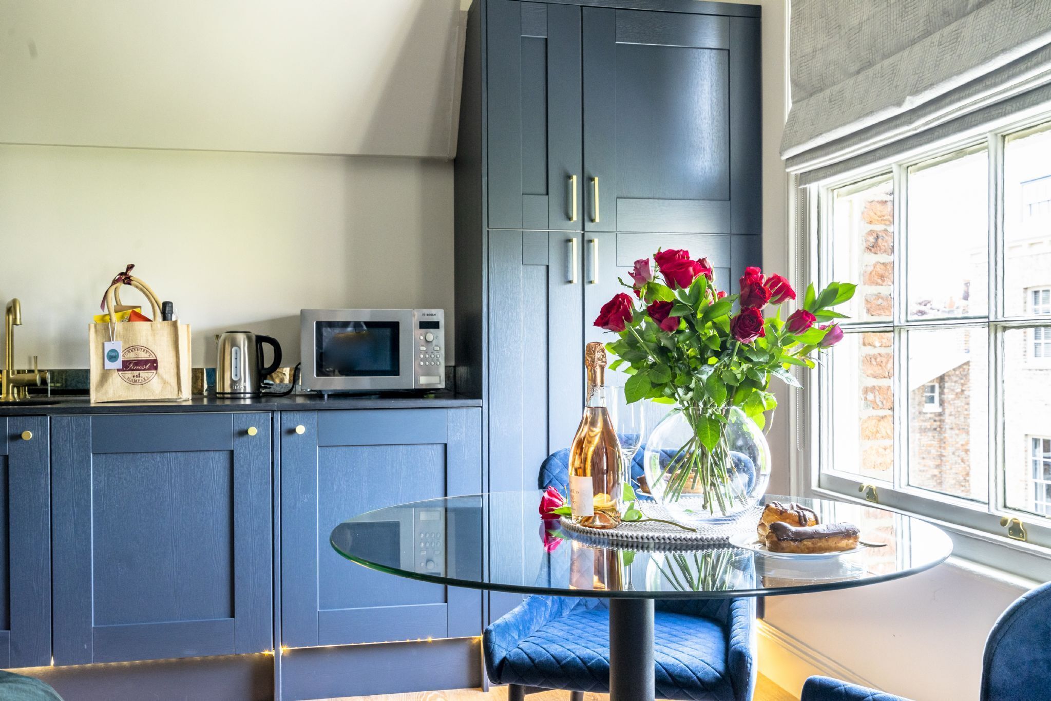 A modern kitchen with dark blue cabinets, a glass-top dining table, and a view of the city through a large window.