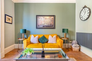 A living room with a yellow sofa, two side tables with lamps, a framed abstract painting, a wall clock, and a glass coffee table with a plant.