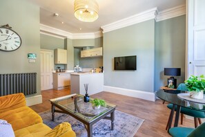 A modern living room with a glass coffee table, a sofa, a dining table with chairs, a kitchen area with cabinets and appliances, and a mounted television.