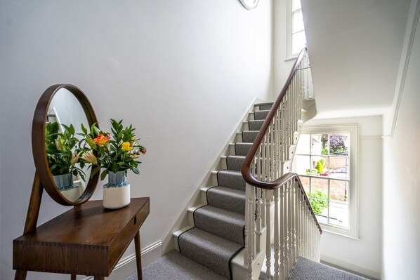 Una escalera con barandilla de madera y escalones alfombrados, una consola de madera con espejo y plantas en macetas, y una ventana con vista al exterior.