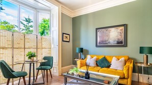 A living room with a yellow sofa, green chairs, a coffee table, and a large window with a view of trees. - A premium stay that sleeps 2 guests  in 1 bedroom (York)