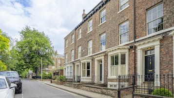 A row of brick townhouses with white window frames, a sidewalk, and a street with parked cars.