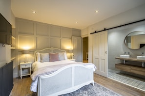 A bedroom with a white bed, bedside lamps, a wooden floor, a large mirror, and a bathroom with a sink and toilet. - A couples retreat that sleeps 2 guests  in 1 bedroom (York)