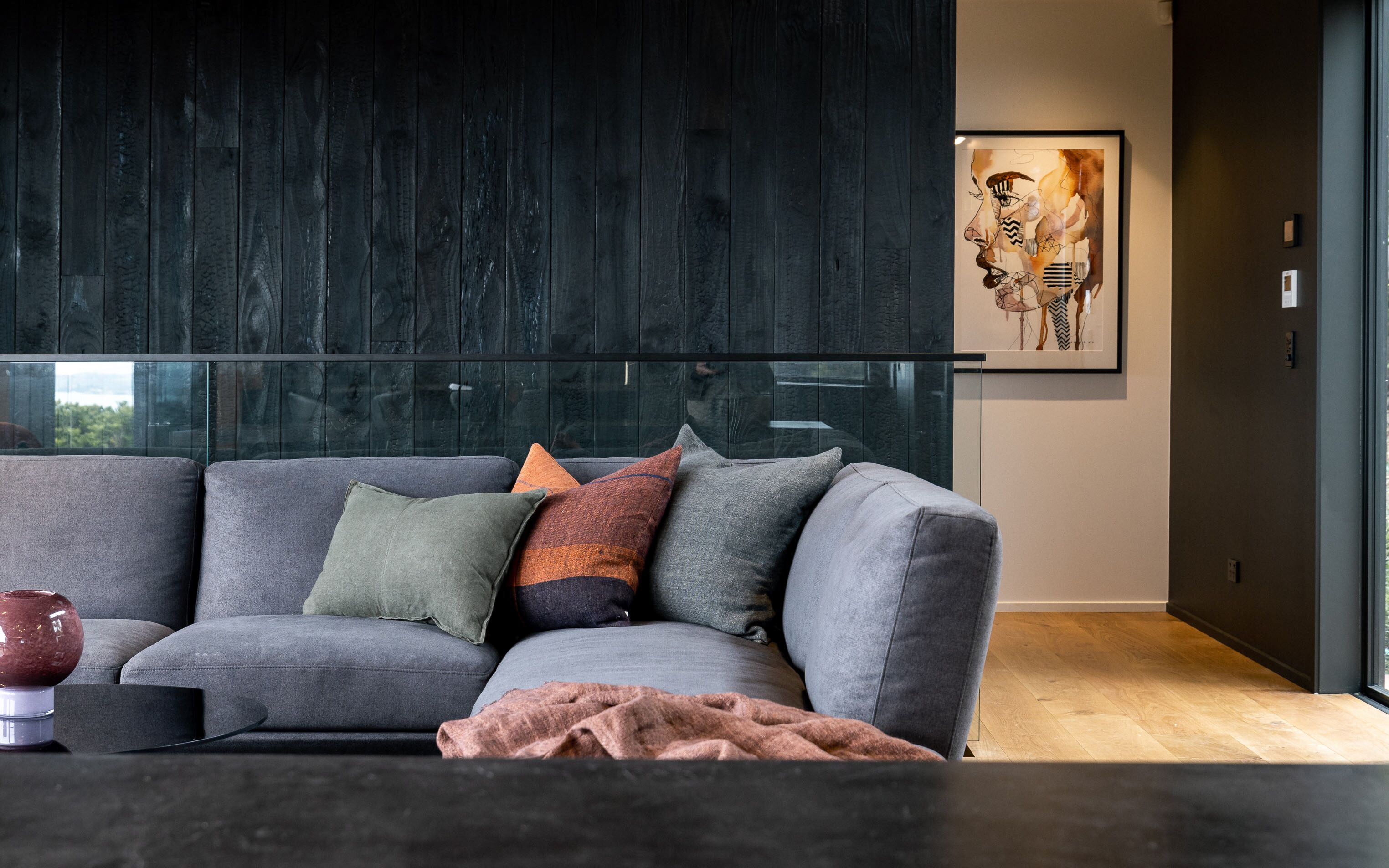 A modern living room with a grey sofa, multiple cushions, a glass coffee table, and a framed artwork on the wall.