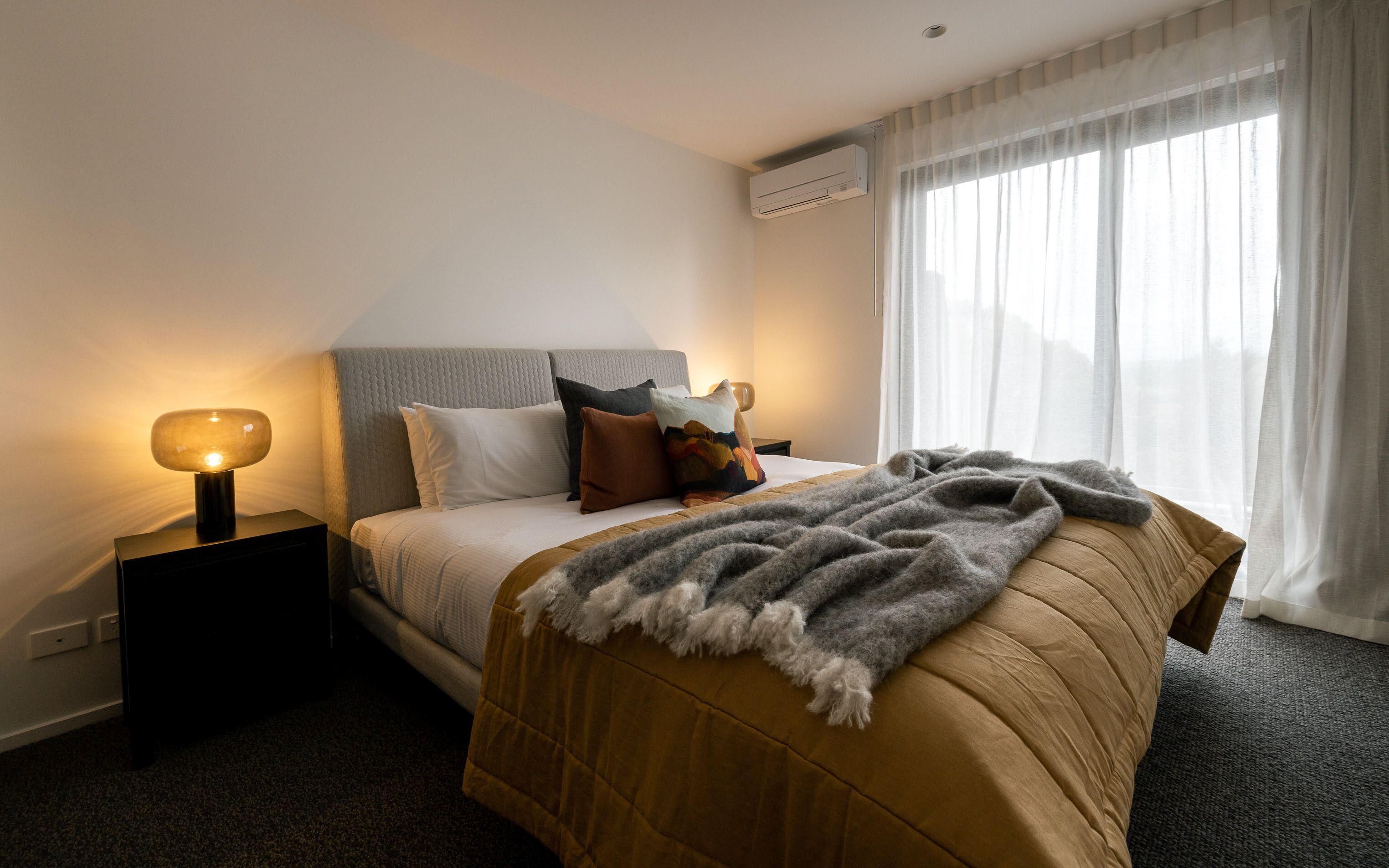 A hotel room with a bed, a bedside lamp, a window with sheer curtains, and an air conditioning unit.
