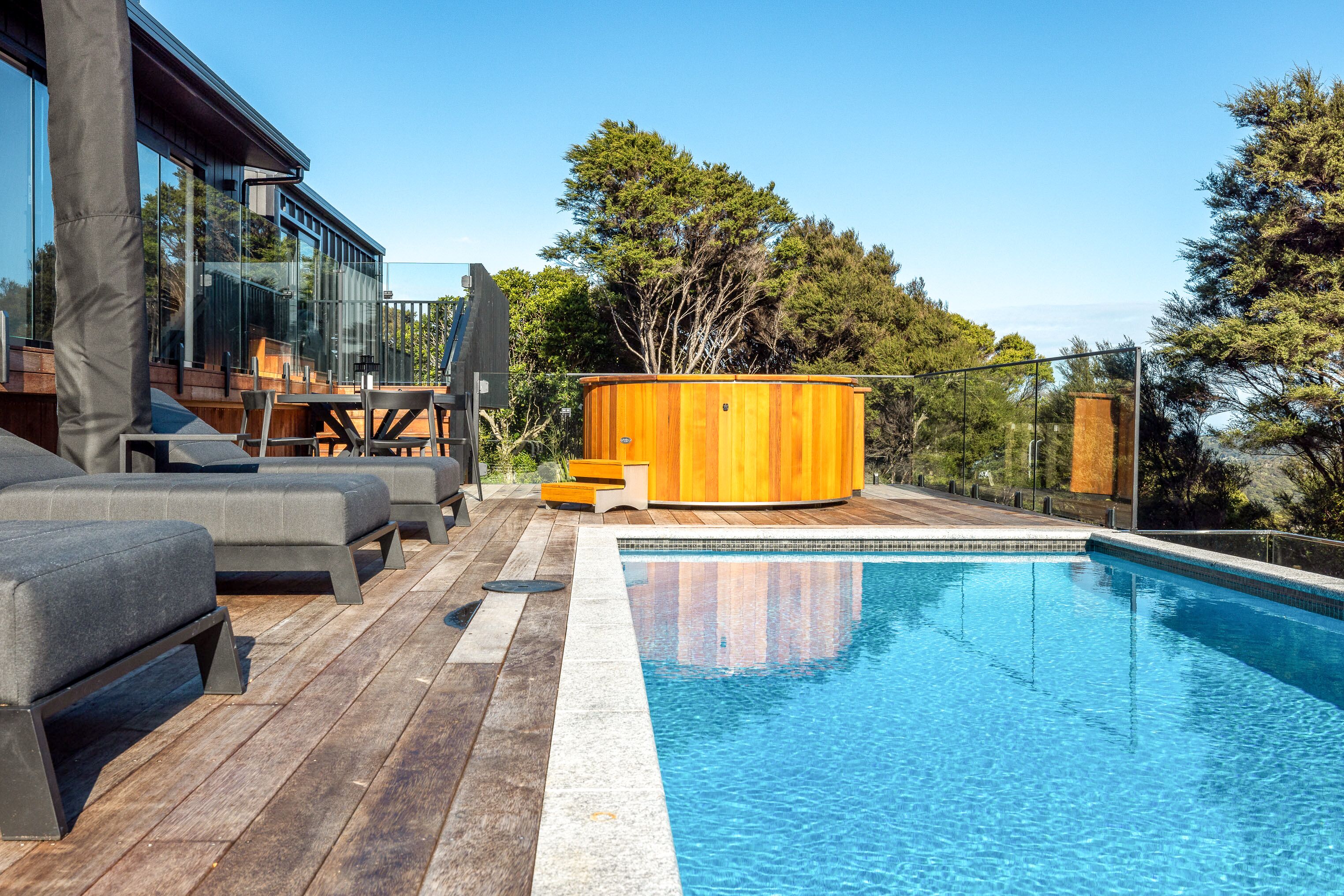 A modern outdoor pool area with wooden deck, lounge chairs, and a hot tub.