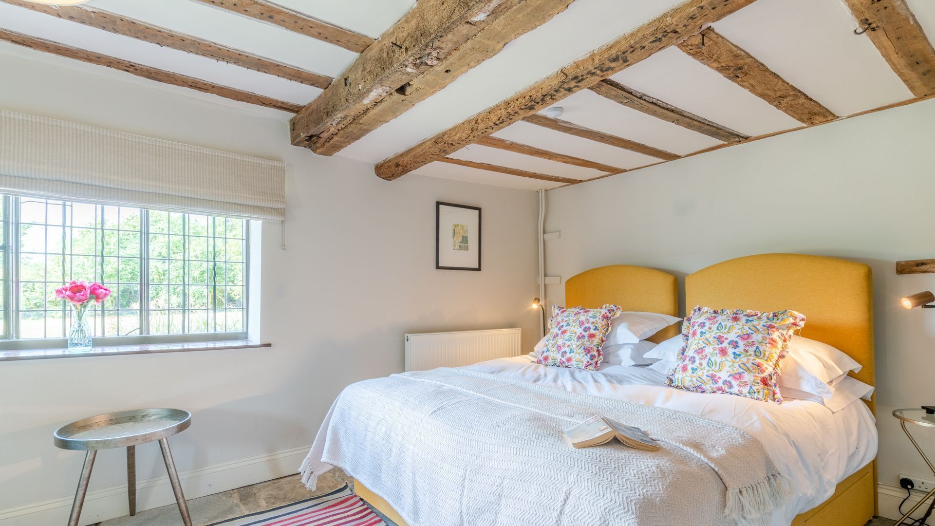 A bedroom with a bed, a window with flowers, a radiator, and a small table.