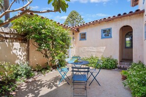 Une terrasse avec une table et des chaises, entourée de plantes et bordée d’un bâtiment dont le toit est recouvert de tuiles.