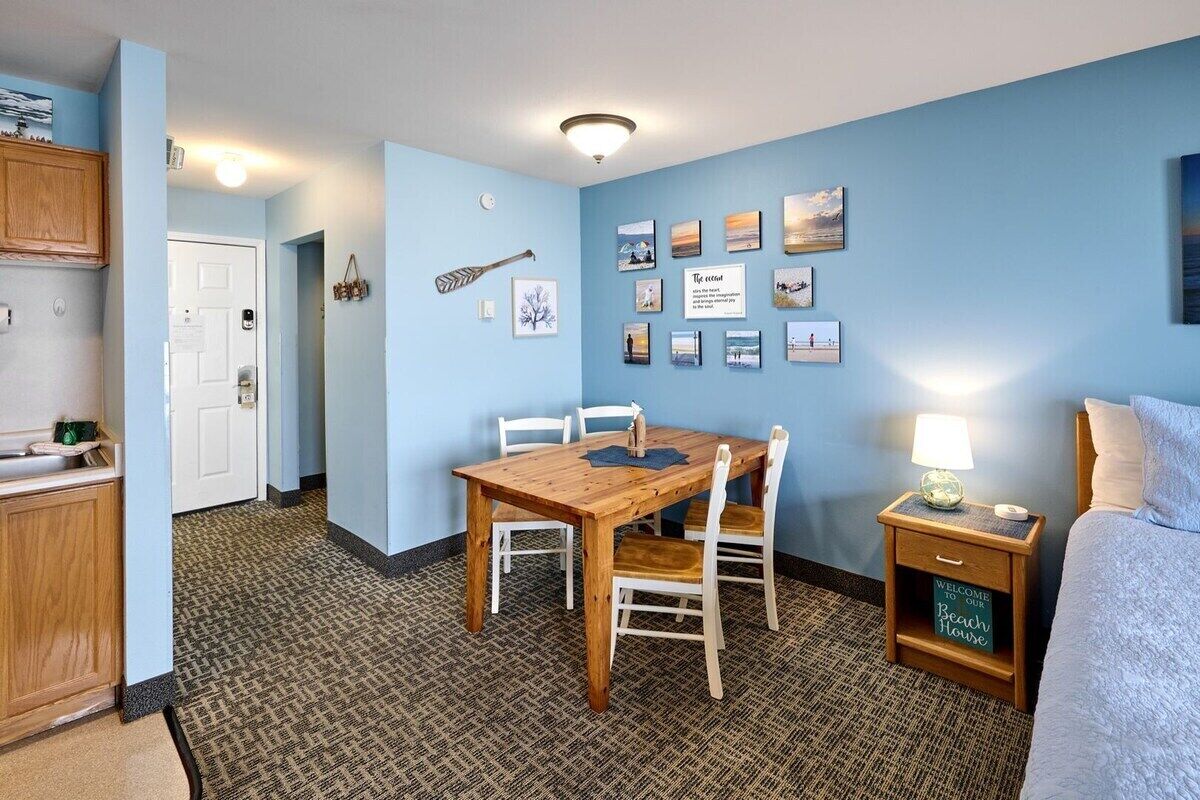 A room with a wooden dining table, white chairs, a bed with a white comforter, a nightstand with a lamp, and a wall with framed pictures.