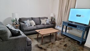 A living room with a grey sofa, a wooden coffee table, a TV on a stand, and a lamp on a side table. - Chateau 16, spacious townhouse in Tickfaw, LA (Tickfaw)