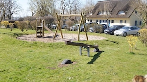 A park with a swing set, a seesaw, and a sandbox. There are cars parked in the background and a house with a thatched roof. - Thatched roof house Hasel 2 (Poseritz)