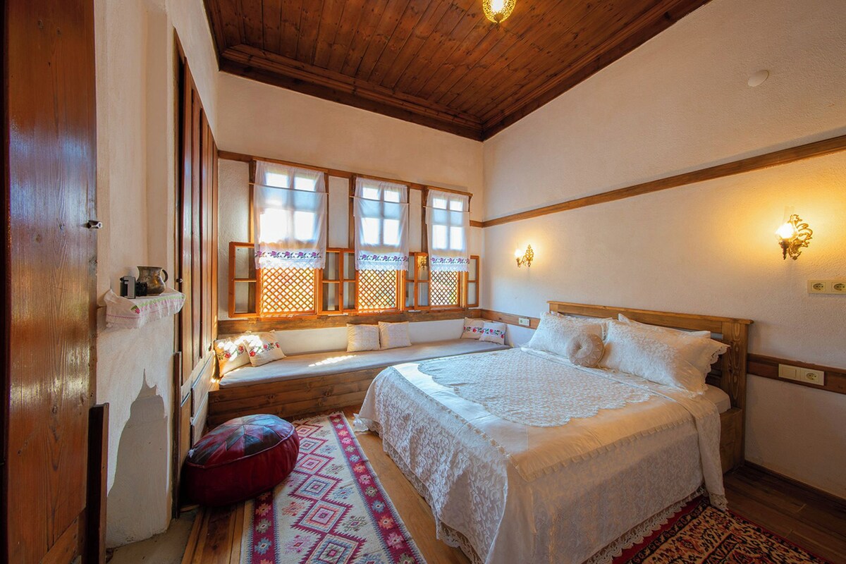A traditional Japanese-style bedroom with a wooden ceiling, tatami mat flooring, a large bed with white bedding, a window seat with curtains, and wall-mounted lighting.