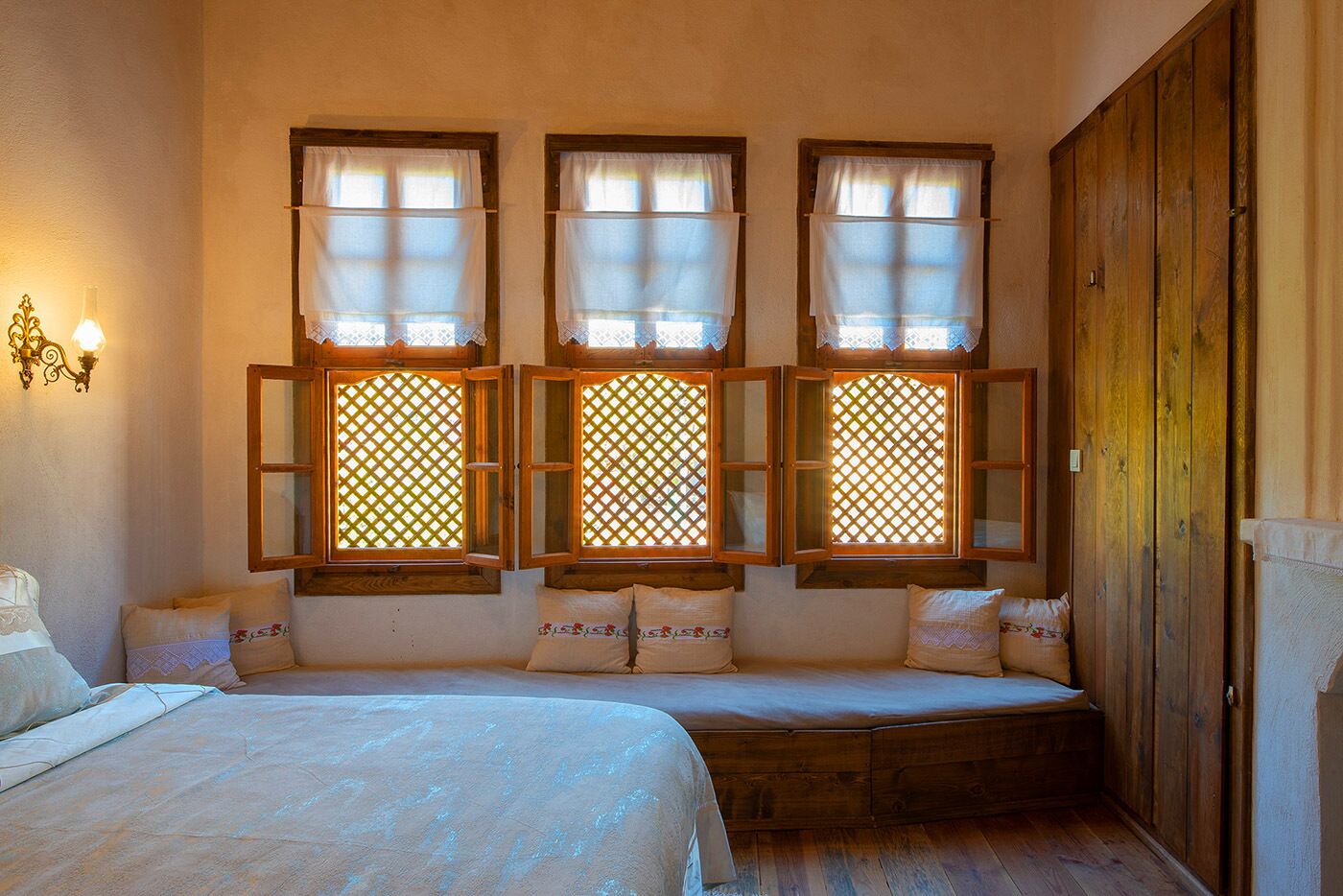 Ein Zimmer mit drei Fenstern mit Gitterornamenten, einem Bett mit weißer Bettdecke und einem Holzschrank.