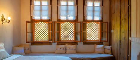 Ein Zimmer mit drei Fenstern mit Gitterornamenten, einem Bett mit weißer Bettdecke und einem Holzschrank.