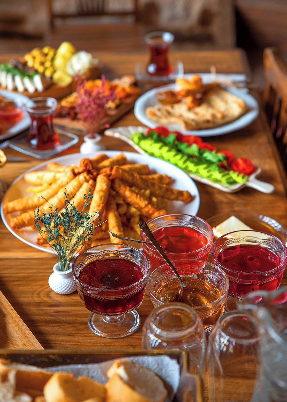 Uma mesa com diversos pratos, incluindo frituras, uma salada e vários copos com líquido vermelho, possivelmente uma bebida ou acompanhamento.