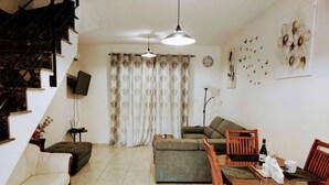 Un saló amb un sofà, una taula de dinar amb cadires, una escala, una televisió i una finestra gran amb cortines.