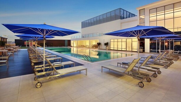 Un área junto a la piscina con sillones de descanso y sombrillas, piscina y un edificio moderno con amplios ventanales.