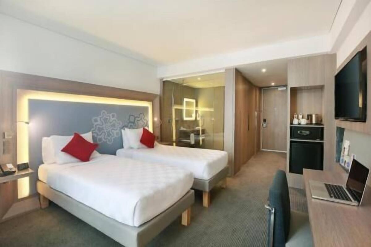 A hotel room with two beds, a desk with a laptop, a TV, and a bathroom visible through a glass partition.
