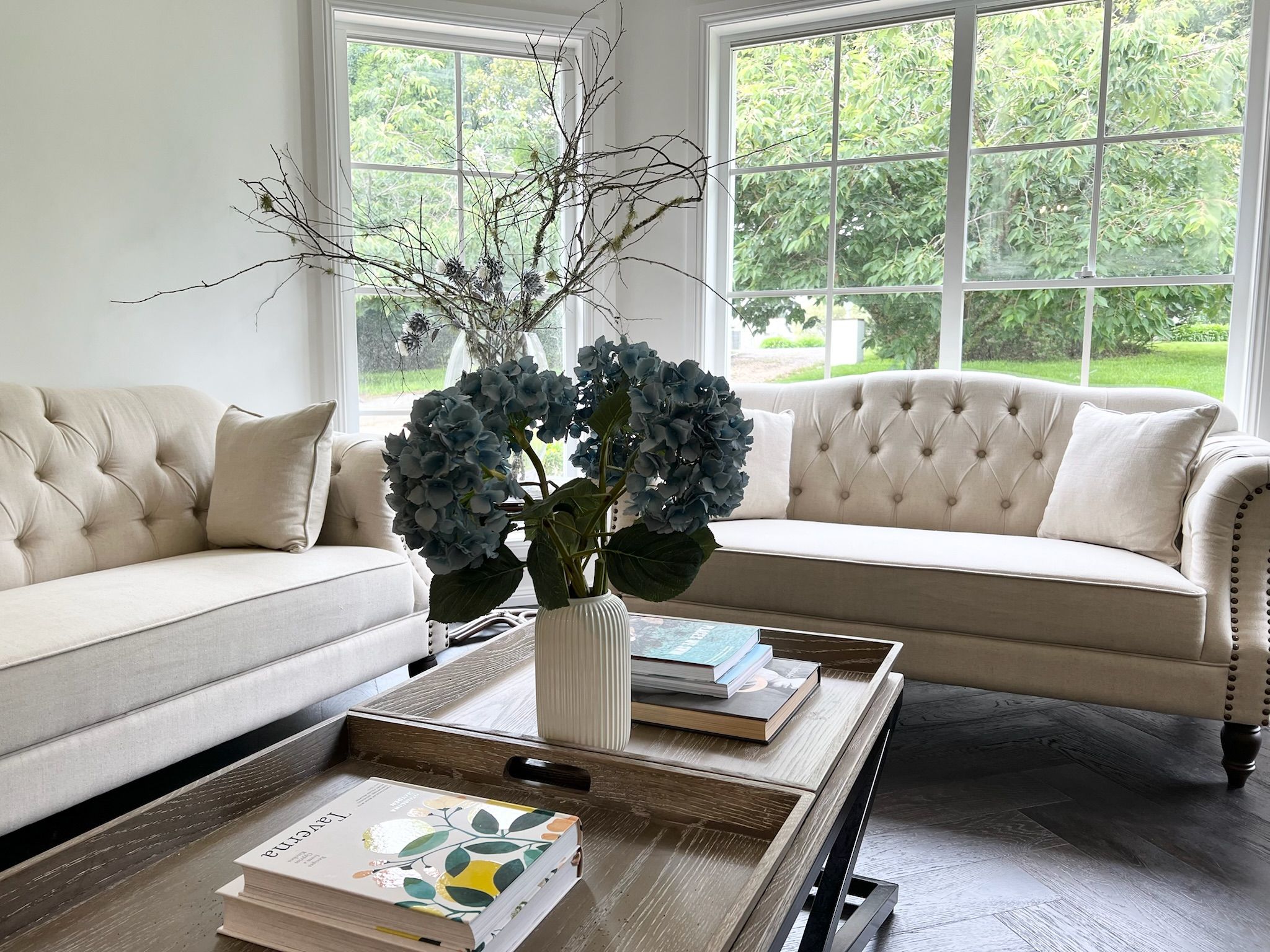 A living room with a beige sofa, a coffee table with books, and a vase with branches and blue flowers.