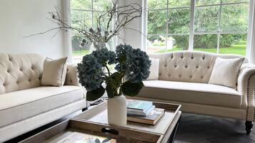 A living room with a beige sofa, a coffee table with books, and a vase with branches and blue flowers.