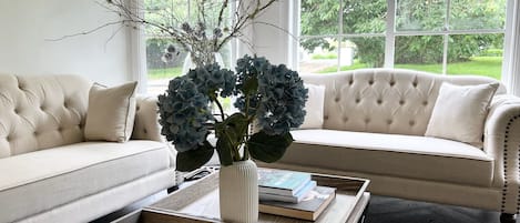 A living room with a beige sofa, a coffee table with books, and a vase with branches and blue flowers.