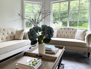 A living room with a beige sofa, a coffee table with books, and a vase with branches and blue flowers.