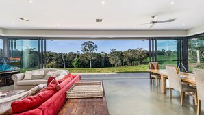 A modern living room with a large glass wall, a wooden dining table, and a red sofa. - The Junction Canyonleigh. Idyllic Weekends (Brayton)