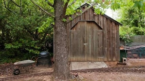 Ein rustikales Holz-Schuppenhaus mit geneigtem Dach, gelegen in einem bewaldeten Gebiet mit einem Baum im Vordergrund und einem Zaun im Hintergrund.